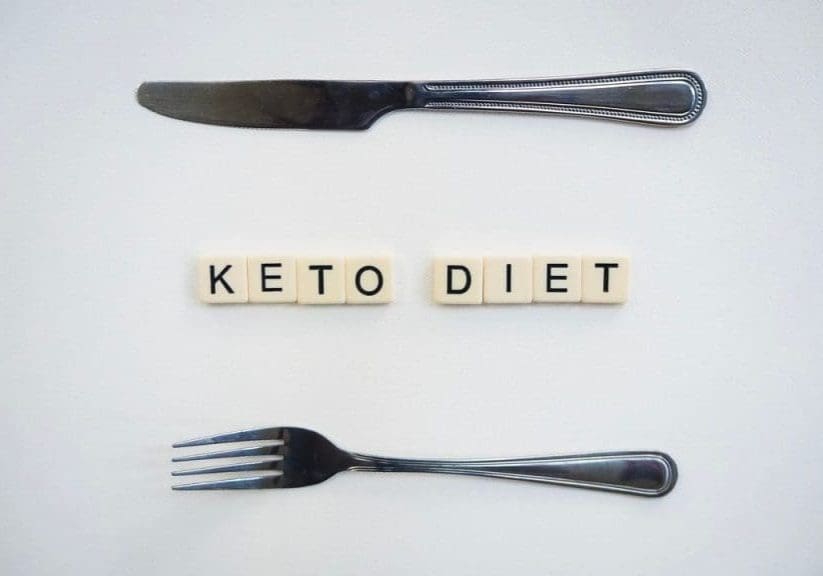 Top view of a knife, fork, and scrabble tiles spelling 'Keto Diet' on a white surface.