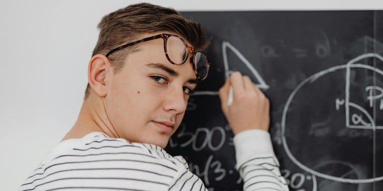 Teen boy writing math equations on a blackboard in a classroom setting.